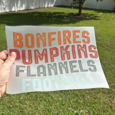 Sheet with text 'Bonfires Pumpkins Flannels Football' held in front of a grassy area.