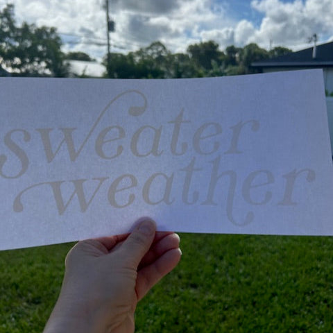 Hand holding a sign with 'sweater weather' text against a cloudy sky.