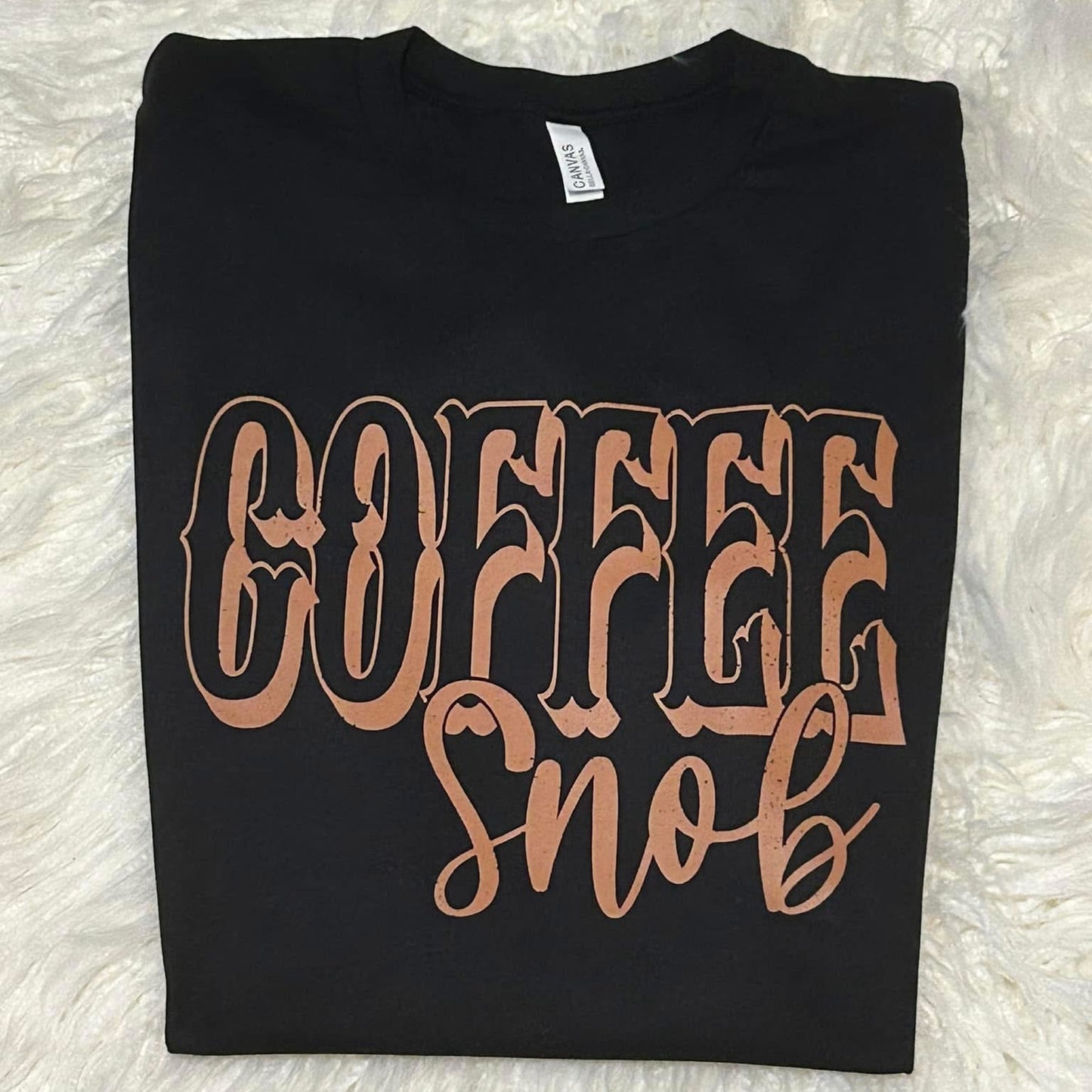 Black t-shirt with 'COFFEE snob' text in rose gold on a white fluffy background