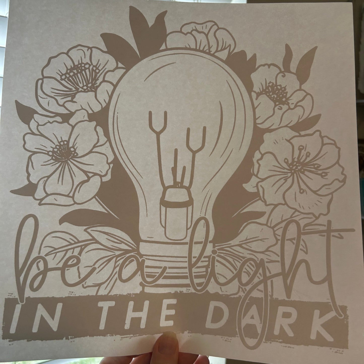 Hand holding a printed sheet with floral design and text 'Light in the Dark'.