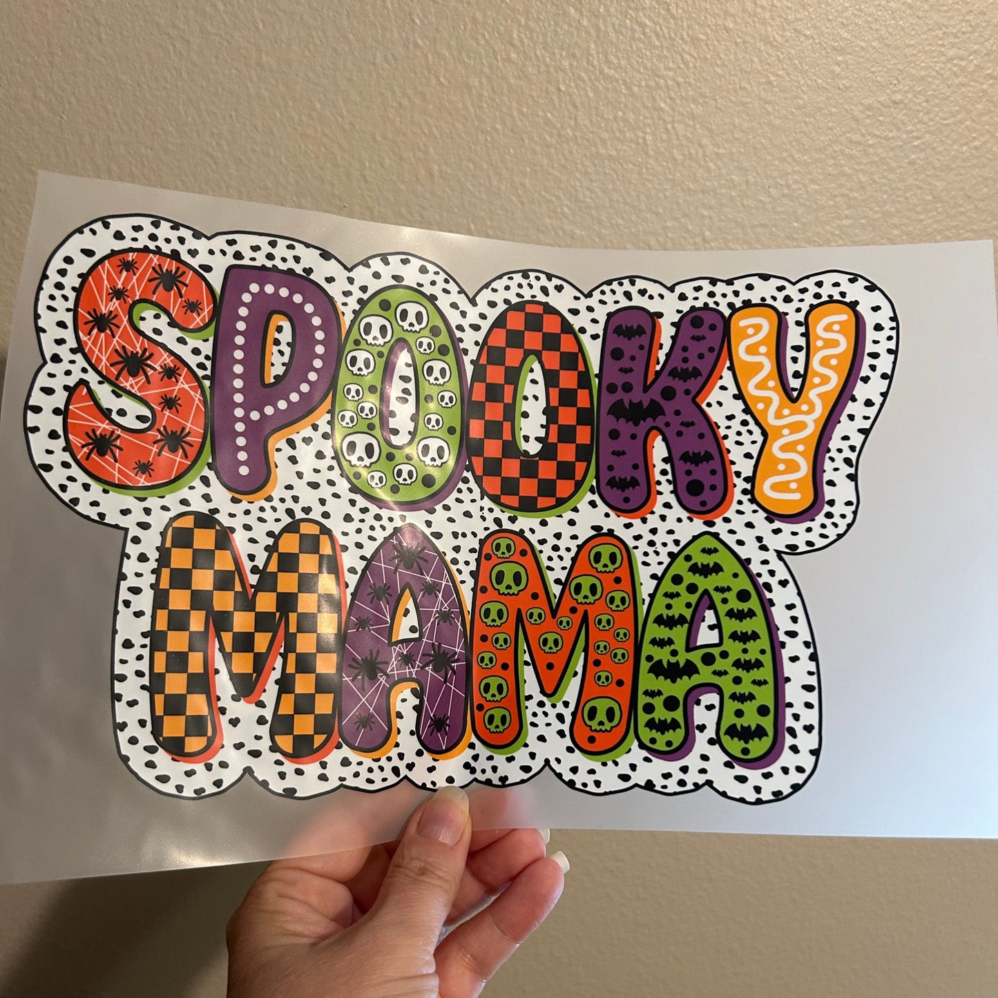 Sheet of Halloween-themed stickers with colorful designs held by a hand against a beige wall.