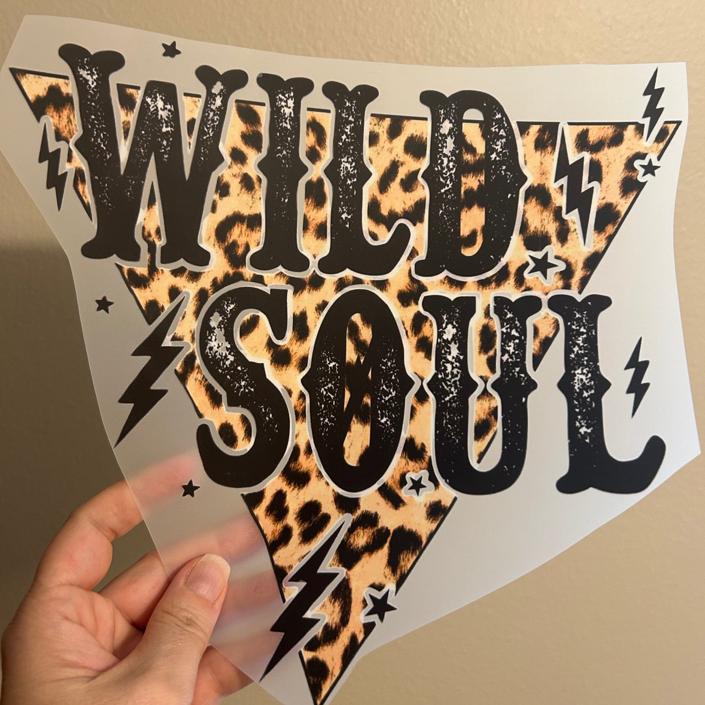 Hand holding a sheet of decorative vinyl with leopard print and black designs on a beige background