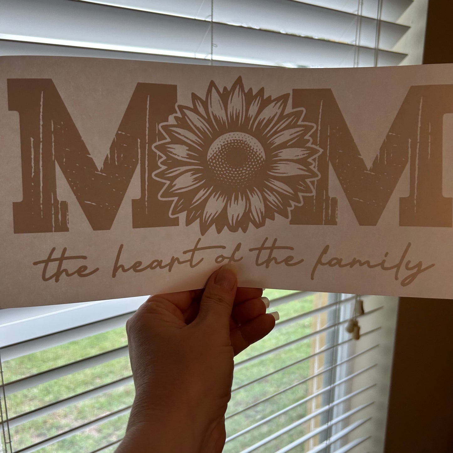 Hand holding a vinyl decal with a sunflower design and text against a window background
