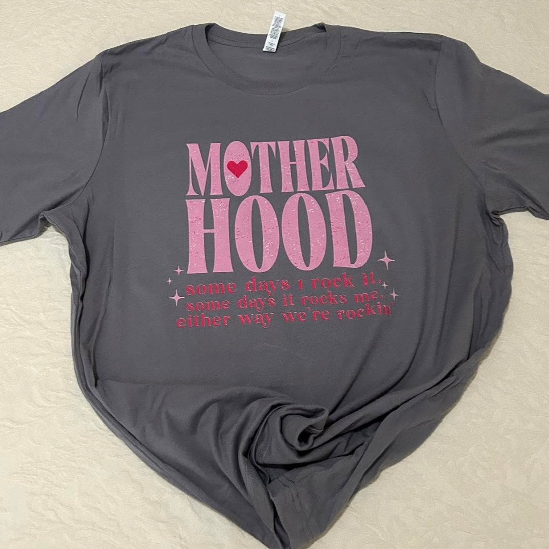 Gray t-shirt with pink 'MOTHER HOOD' text on a beige surface