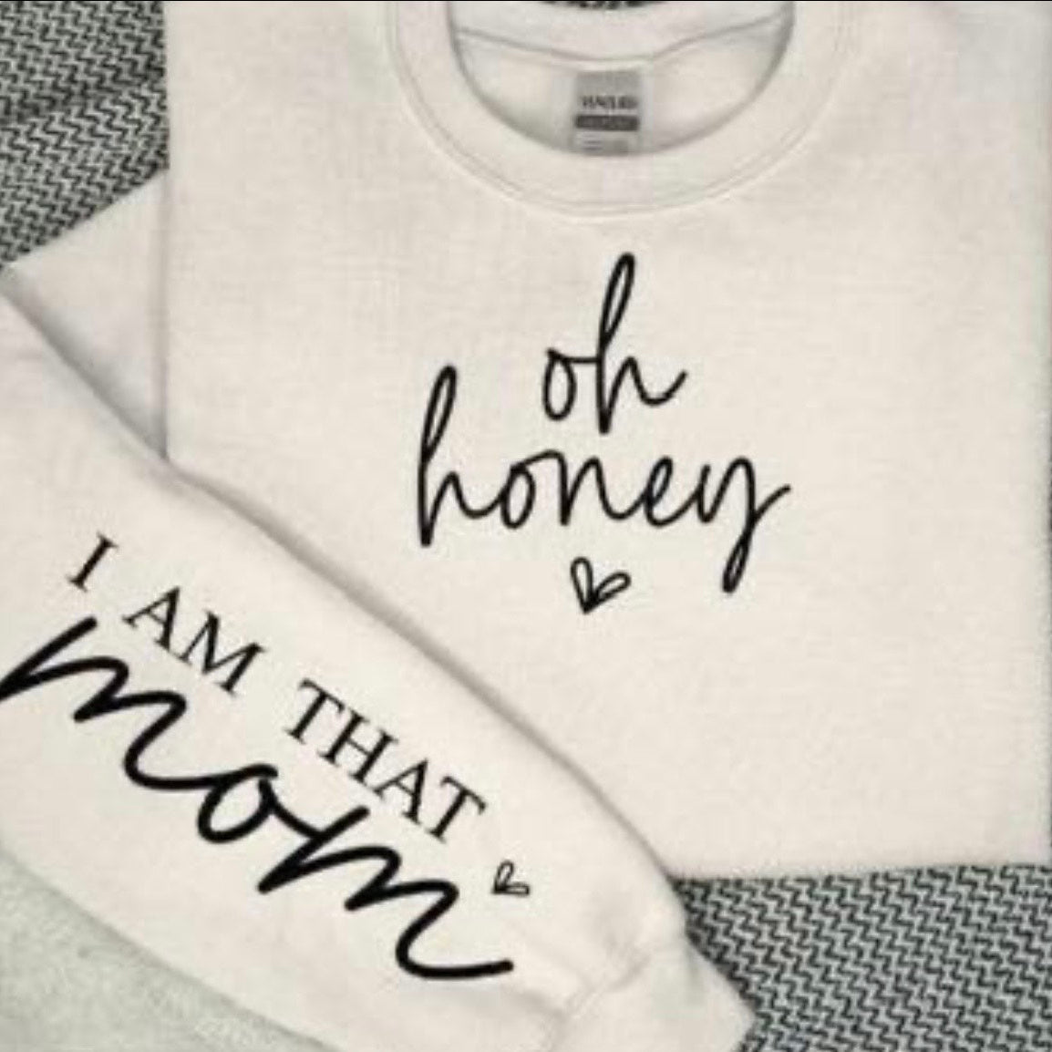 White sweatshirt with text 'oh honey I am that mom' on a textured surface